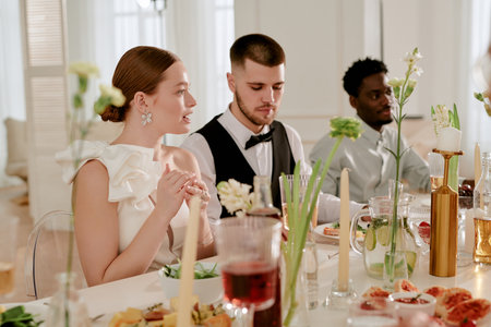Caucasian young adult woman sitting beside Caucasian young adult man and Black young adult man at wedding celebration table, engaging in conversation and enjoying festive meal togetherの写真素材