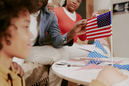 Child sitting at table with mom and dad assembling American flags, dad holding small flag on stick,...