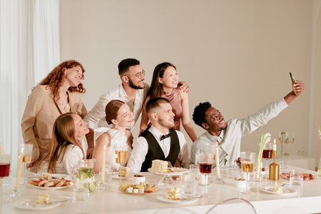 Group of young adult multiethnic men and women smiling and posing for selfie at wedding celebration, sitting and standing around decorated banquet table with food and drinksの写真素材