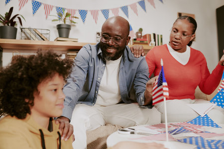 Black middle aged man and Black middle aged woman sitting together smiling and talking with Black child holding American flag, celebrating patriotic holiday indoors with decorationsの写真素材