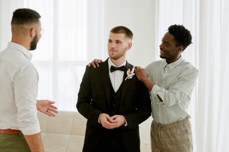 Caucasian young adult man in tuxedo standing between Black young adult man and Caucasian young adult man, friends supporting and talking during wedding celebration indoorsの写真素材