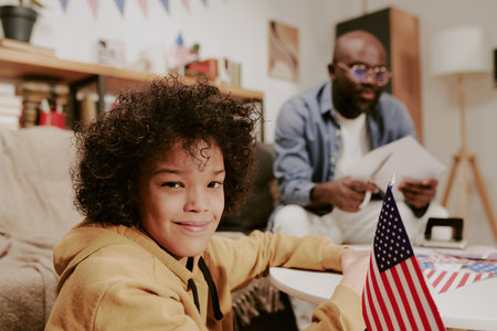 Portrait of smiling child sitting at table holding American flag, looking into camera while middle aged man reading documents in background, home interior visibleの写真素材