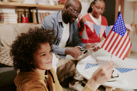 Child holding American flag while smiling, sitting at table with Black middle aged man and Black woman cutting paper flags, celebrating patriotic holiday indoorsの写真素材