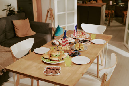 Table set with burgers, cupcakes, cookies, fruit, corn on cob, drinks and two American flags, suggesting festive meal preparation for holiday celebration in modern home dining areaの写真素材