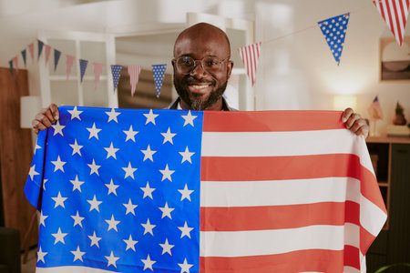 Portrait of Black middle aged man smiling while holding large United States flag indoors, American flag bunting hanging in background, celebrating national holiday or patriotic eventの写真素材