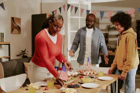 Black middle aged woman arranging food on table with American flag while Black middle aged man and Black teenager boy standing nearby, celebrating holiday with festive meal indoorsの写真素材