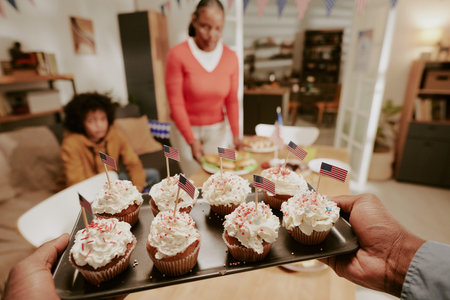 Male hands presenting tray of cupcakes decorated with American flags in foreground, Black middle aged woman and Black teenager sitting at table in background during celebrationの写真素材