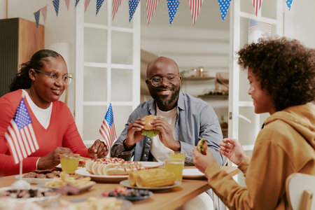Black middle aged man and Black middle aged woman sitting at table with Black teenage boy, eating burgers and hot dogs, holding American flags, celebrating together indoorsの写真素材