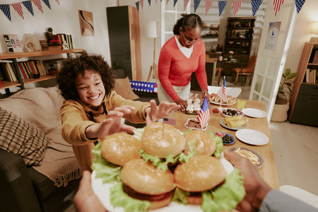 Black woman arranging desserts on table while Black child reaching excitedly for tray of hamburgers during festive indoor celebration with American flagsの写真素材