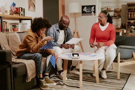 Teenage boy sitting on sofa assembling American flags with father and mother smiling and interacting...