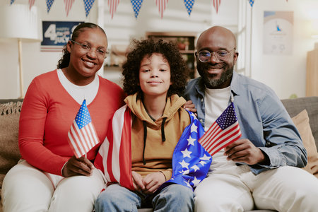 African American middle aged woman and man sitting with teenage boy holding American flags and draped in United States flag, smiling together on couch during patriotic celebrationの写真素材
