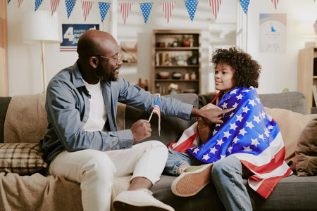 African American dad handing small American flag to child wrapped in United States flag, both sitting on sofa, celebrating patriotic holiday with festive decorationsの写真素材