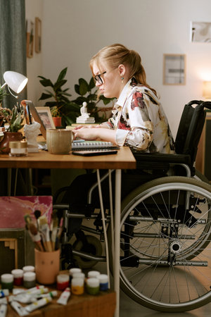 Caucasian young adult woman with disability sitting in wheelchair working at desk using laptop and...