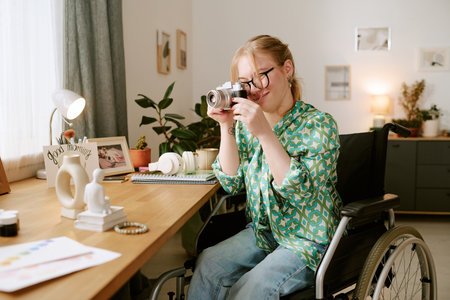 Caucasian young adult woman with disability sitting in wheelchair holding camera and taking photo at...