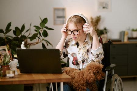 Caucasian young adult woman with disability sitting in wheelchair using laptop and wearing headphones, small brown dog resting on her lap, working or studying at homeの写真素材