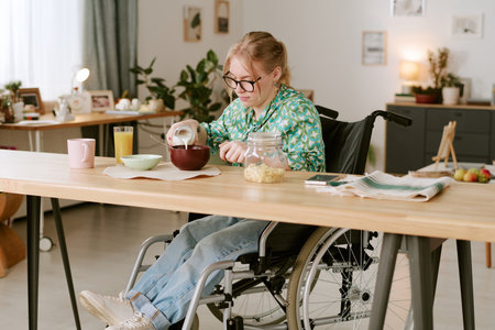 Caucasian woman with disability sitting in wheelchair pouring milk into bowl at kitchen table eating breakfast with glass of juice, cookies in jar, newspaper and smartphone nearbyの写真素材
