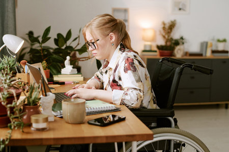 Caucasian young adult woman with disability sitting in wheelchair working on laptop at desk...