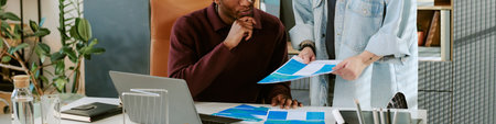 Black man sitting at desk analyzing documents while young adult Caucasian woman standing beside him holding charts, both collaborating on business project in modern office workspaceの写真素材