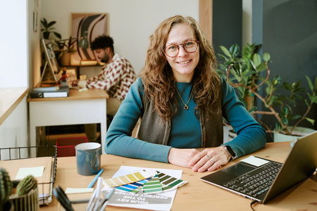 Portrait of middle aged Caucasian woman with long curly hair smiling at camera while sitting at desk...