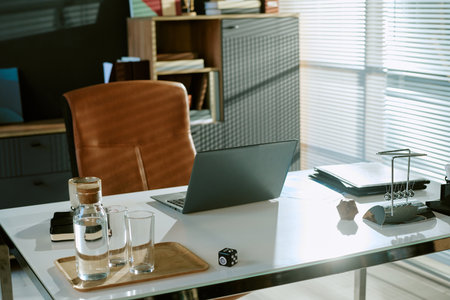 Modern office workspace featuring closed laptop, glasses of water, coffee cup, documents, and office...