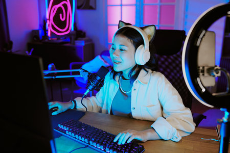 Teenage girl wearing headphones sitting at desk live streaming video games and speaking into microphone with computer monitor and ring lightの写真素材