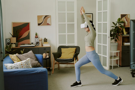 Teenage Caucasian girl wearing VR headset exercising in living room, performing yoga pose with arms raised, engaging in virtual reality fitness activity at homeの写真素材
