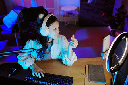 Teenage girl sitting at desk wearing headphones and speaking into microphone while live streaming video gamesの写真素材