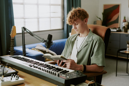 Teenage boy sitting at desk reading book while recording music with microphone and digital keyboard...