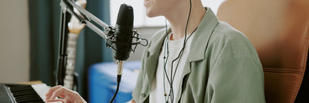 Website banner of teenage boy singing into microphone while playing keyboard, wearing earphones, recording music at home, close up of face and hands, guitar visible in backgroundの写真素材