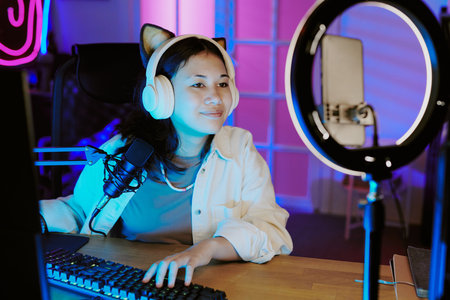 Teenage girl sitting at desk live streaming video games, wearing headphones with cat ears, speaking...