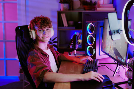 Portrait of teenage boy wearing headphones sitting at gaming desk live streaming video games with...