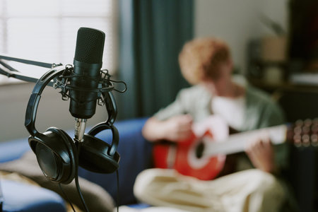 Teenage boy sitting on sofa playing acoustic guitar in home setting, professional microphone and...
