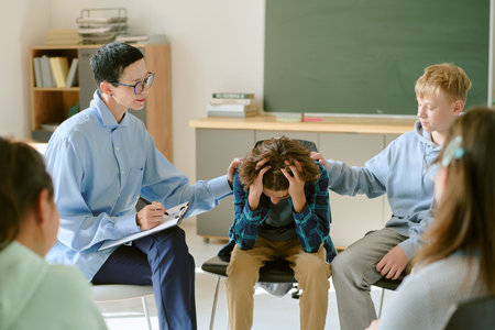 Teenage boy sitting with head in hands being comforted by teenage boy and adult woman holding clipboard during group therapy sessionの写真素材