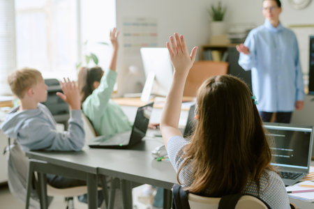 Group of teenagers sitting at desks raising hands while using laptops in classroom, teacher standing in front of students engaging with class activityの写真素材