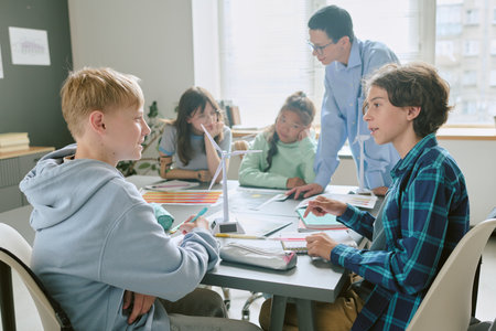 Group of multiethnic teenagers collaborating on school project at desk while teacher supervising and assisting, students engaging in teamwork and discussion in classroom settingの写真素材