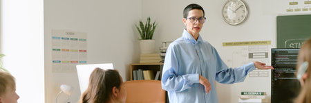 Website banner of middle aged Caucasian woman teaching group of teenagers in classroom, gesturing toward computer monitor displaying code, students listening attentively, educational setting visibleの写真素材