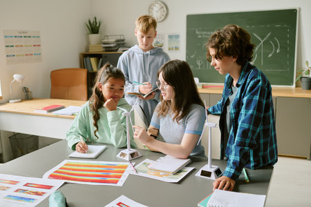 Group of diverse teenagers collaborating on science project in classroom, girl explaining wind turbine model while other teenagers listening and taking notesの写真素材