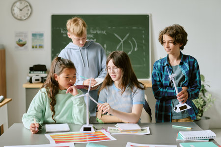 Group of multiethnic teenagers collaborating on science project, assembling wind turbine models and discussing renewable energy concepts in classroom with educational materials visibleの写真素材