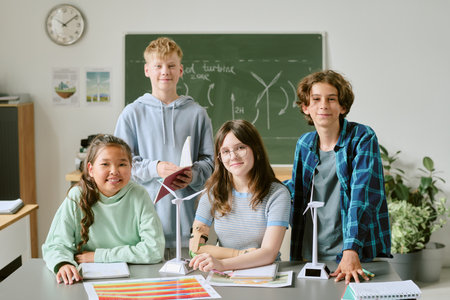 Group of multiethnic teenagers working on wind turbine models in classroom, standing and sitting at desk with charts and notebooks, smiling and looking at cameraの写真素材