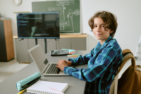 Portrait of teenage boy sitting at desk using laptop and looking at camera in classroom setting with notebook and pen case on table, digital screen in backgroundの写真素材