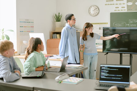Teenage boy and teenage girl standing near interactive display, girl pointing at screen while two...