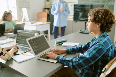 Teenage Caucasian boy working on laptop with code on screen in classroom, surrounded by classmates including Asian girl and student with prosthetic arm, teacher standing near chalkboardの写真素材