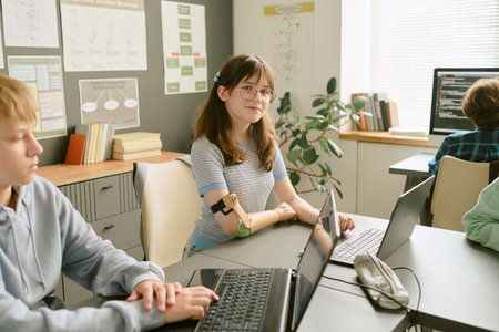 Portrait of Caucasian teenage girl with prosthetic arm sitting at desk in classroom, looking into camera, surrounded by classmates using laptops, educational posters on wallの写真素材