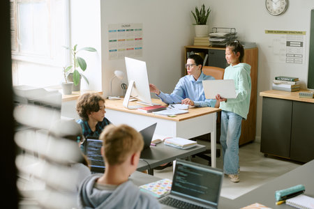 Group of teenagers working together in modern classroom, teacher explaining project on computer to...