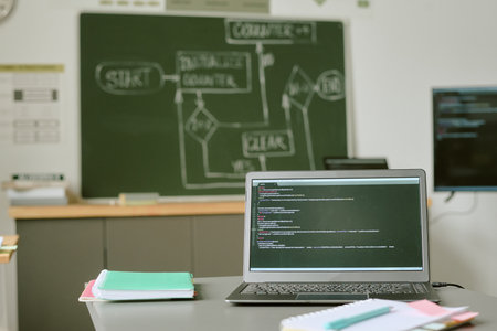 Open laptop displaying programming code on desk in classroom with green chalkboard showing flowchart in background, notebooks and folders scattered on table, no people visibleの写真素材