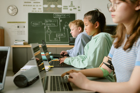 Teenage Caucasian boy, Asian girl, and Caucasian girl with prosthetic arm sitting at desks using laptops in classroom, focusing on tasks, chalkboard with diagrams in backgroundの写真素材