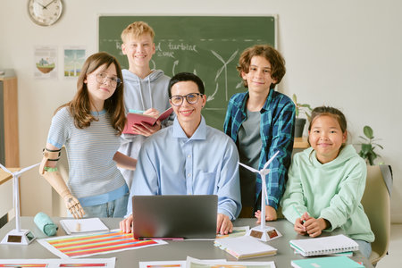 Portrait of teacher with glasses sitting at desk surrounded by diverse group of teenagers smiling and standing in classroomの写真素材
