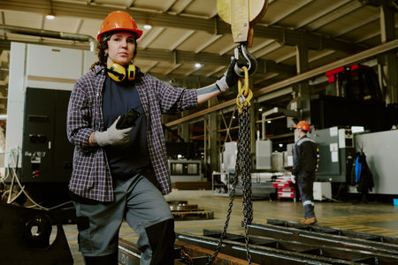 Caucasian young adult woman standing in industrial factory holding radio and crane hook, wearing...