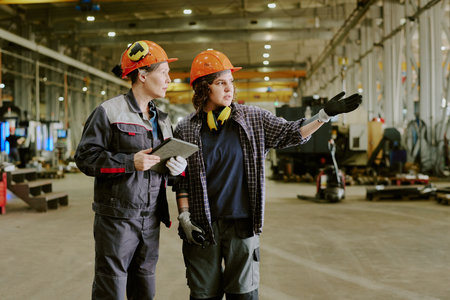 Middle aged woman and young adult woman wearing safety helmets standing in industrial factory,...