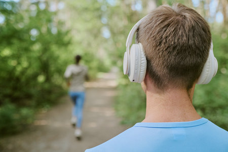 Back view of young Caucasian man wearing headphones standing on forest path watching young woman...